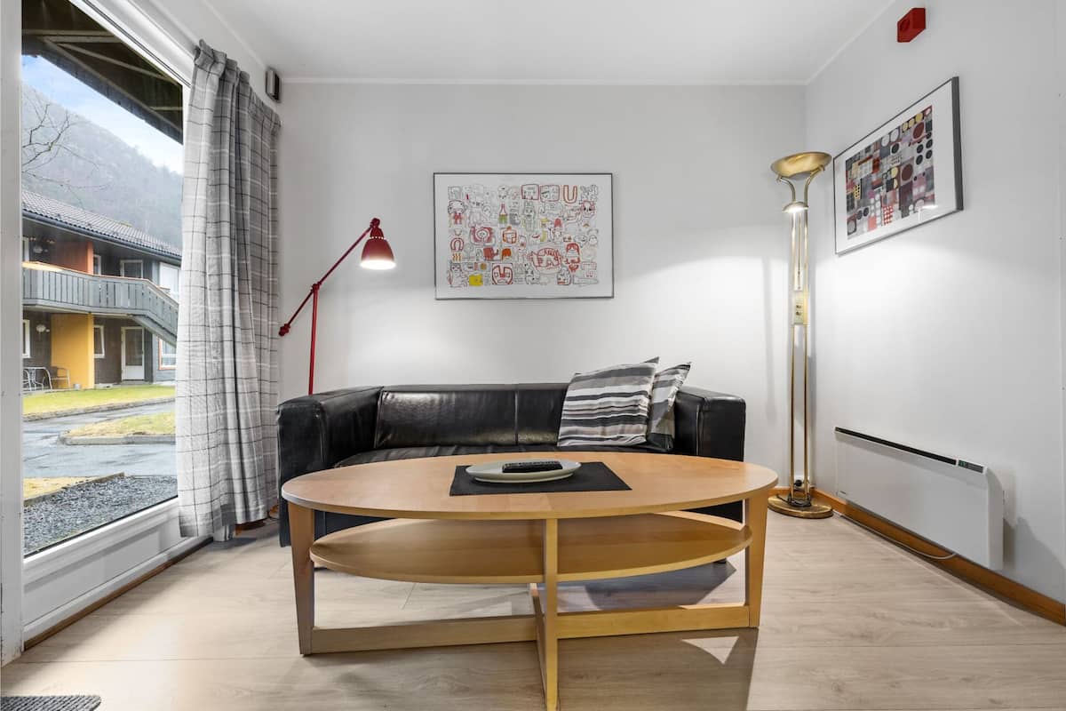 A modern living room with a round wooden coffee table, a black leather sofa, a red floor lamp, and a large window with a view of a building and trees.