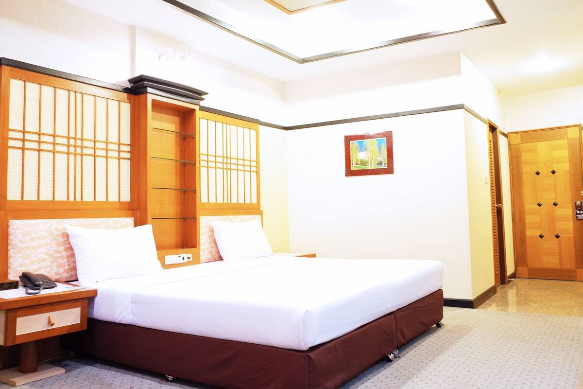 A hotel room with a large bed, a nightstand, a wooden wardrobe, a small table, and a picture on the wall.