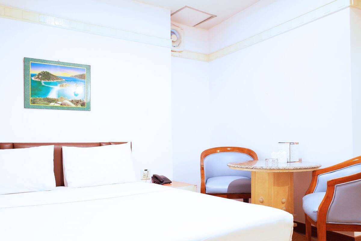 A hotel room with a bed, a desk, two chairs, a painting on the wall, and a small table with a lamp.