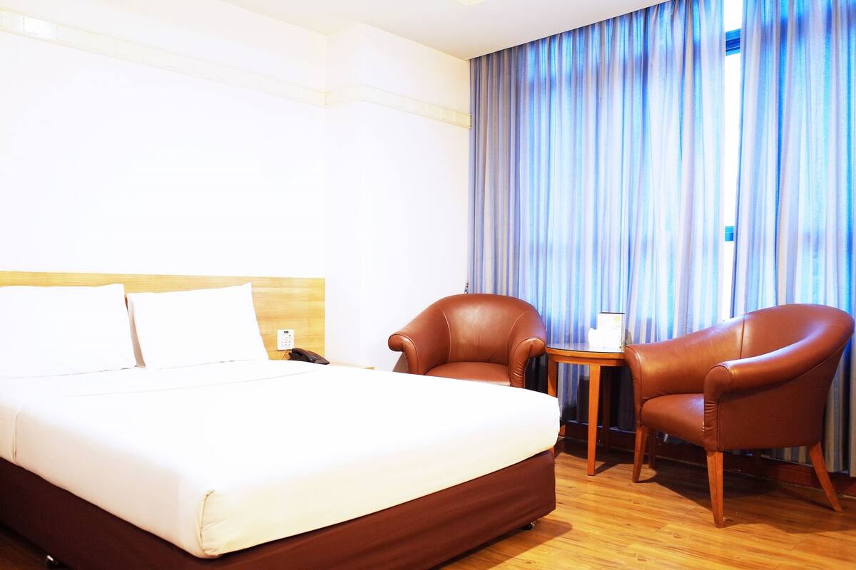 A hotel room with a large bed, two leather armchairs, a small table, and a window with blue curtains.
