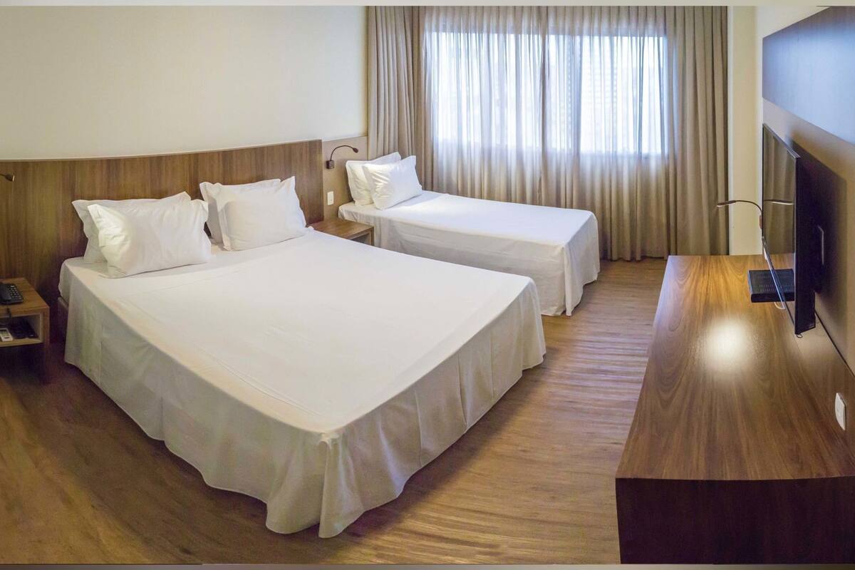 A hotel room with two beds, a wooden nightstand with a phone, a desk with a TV, and a window with curtains.