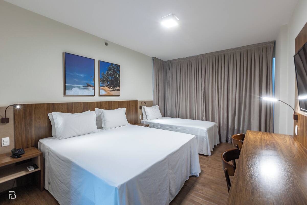 A hotel room with two beds, a wooden desk, a chair, and a large window with curtains.