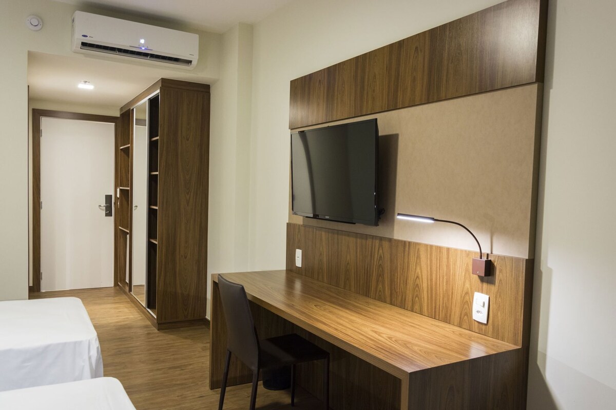 A hotel room with a wooden desk, a television mounted on the wall, a wardrobe, and a bed.