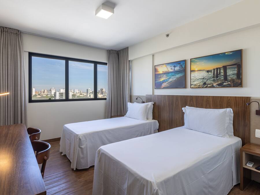 A hotel room with two beds, a wooden desk, a view of the city, and two paintings on the wall.