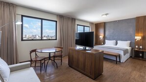 A modern hotel room with a large window, a flat-screen TV, a bed, a wooden desk, and a dining area. - Gran Executive Hotel (Uberlandia)