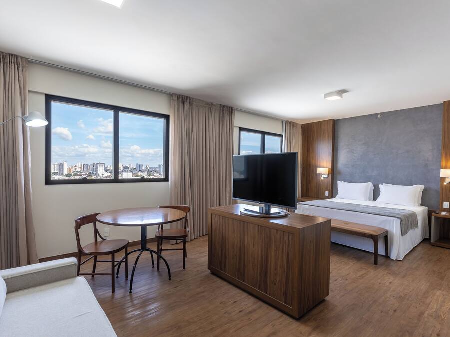 Habitación de hotel moderna con un ventanal grande, televisión de pantalla plana, cama, escritorio de madera y zona de comedor.