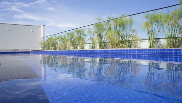 A swimming pool with blue tiles and a clear reflection, surrounded by tall plants and a building in the background. - Gran Executive Hotel (Uberlandia)