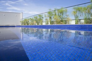 A swimming pool with blue tiles and a clear reflection, surrounded by tall plants and a building in the background. - Gran Executive Hotel (Uberlandia)