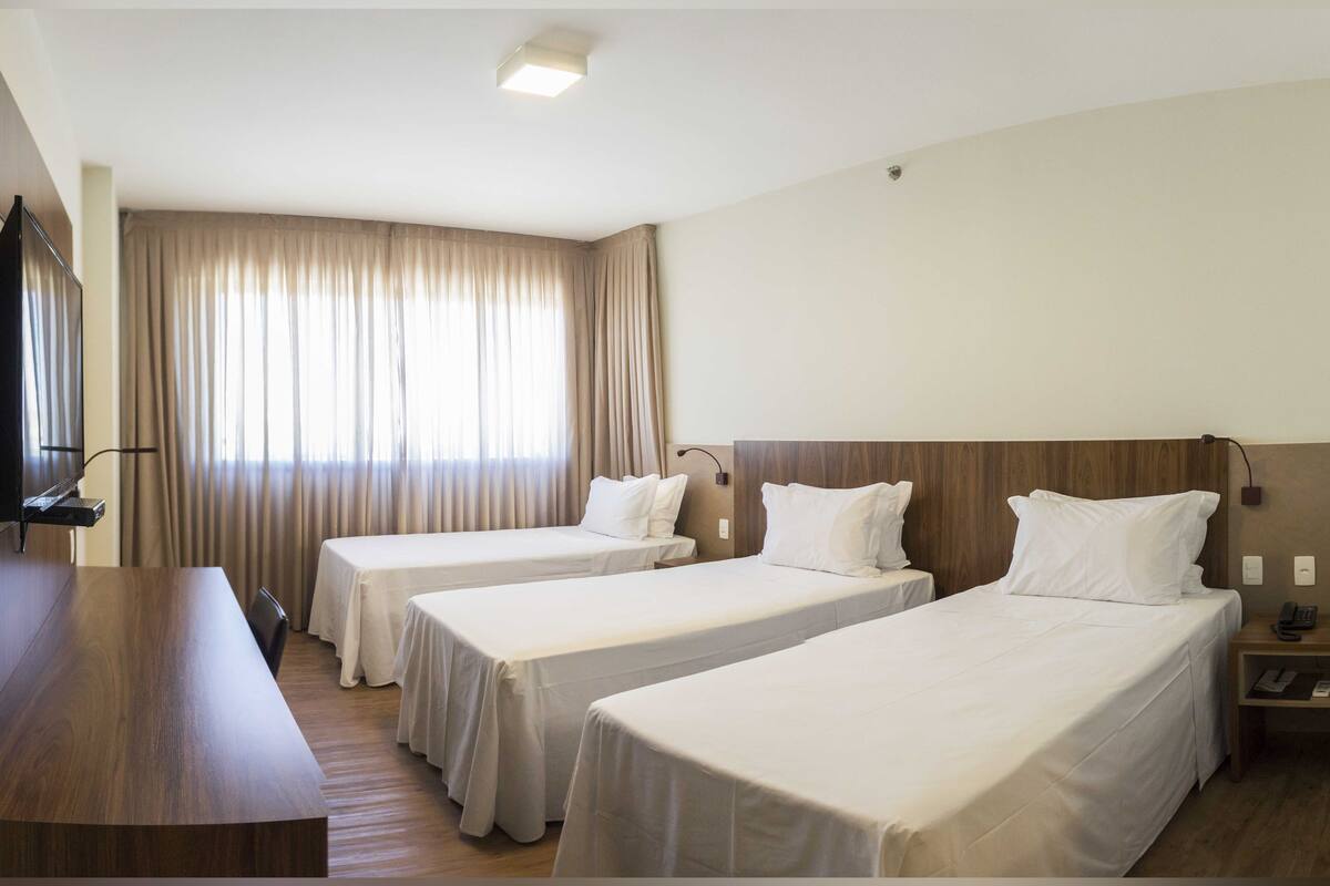 A hotel room with two single beds, a desk with a flat-screen TV, a wooden headboard, and a large window with curtains.