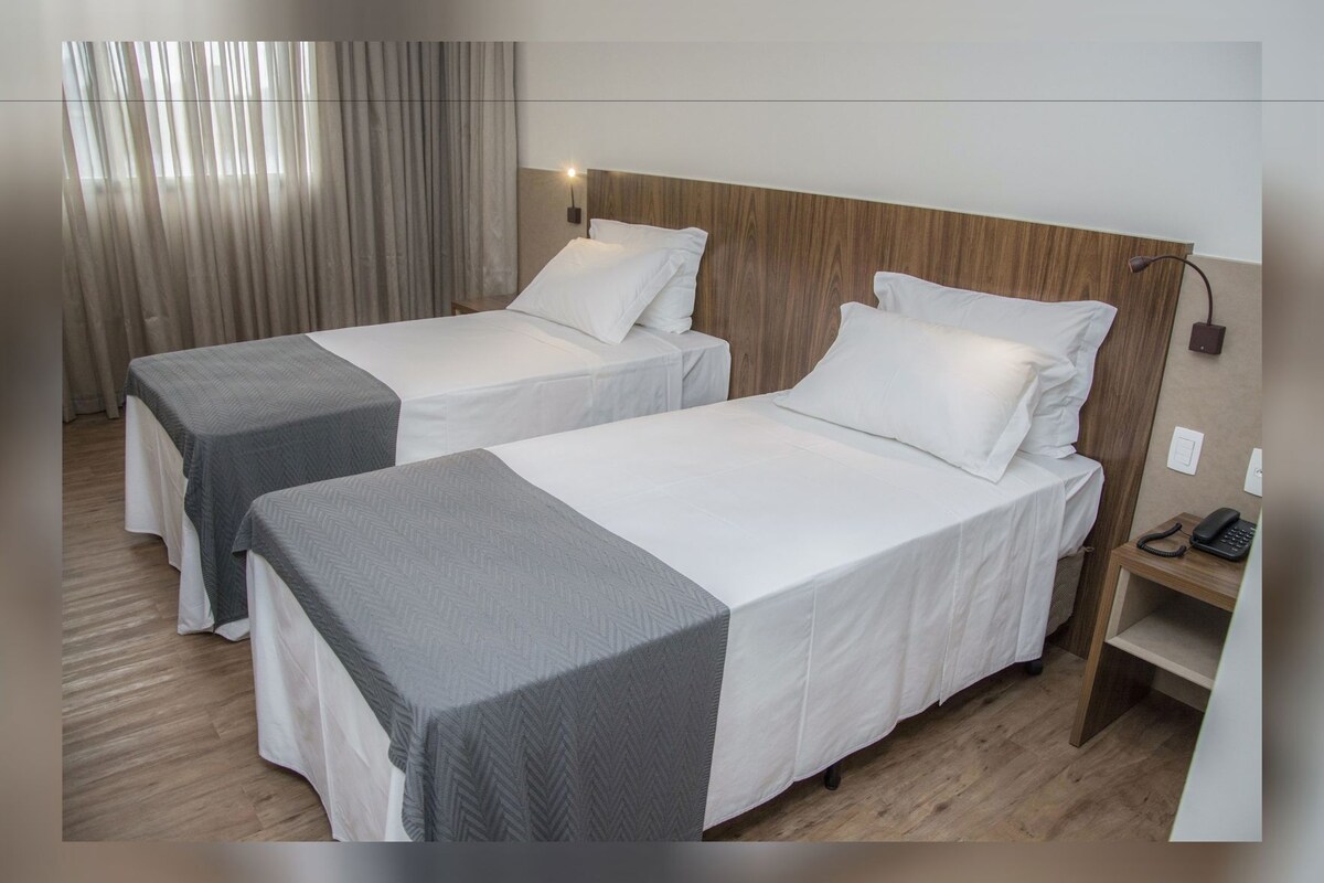 A hotel room with two single beds, a wooden headboard, a bedside table with a phone, and a curtain.