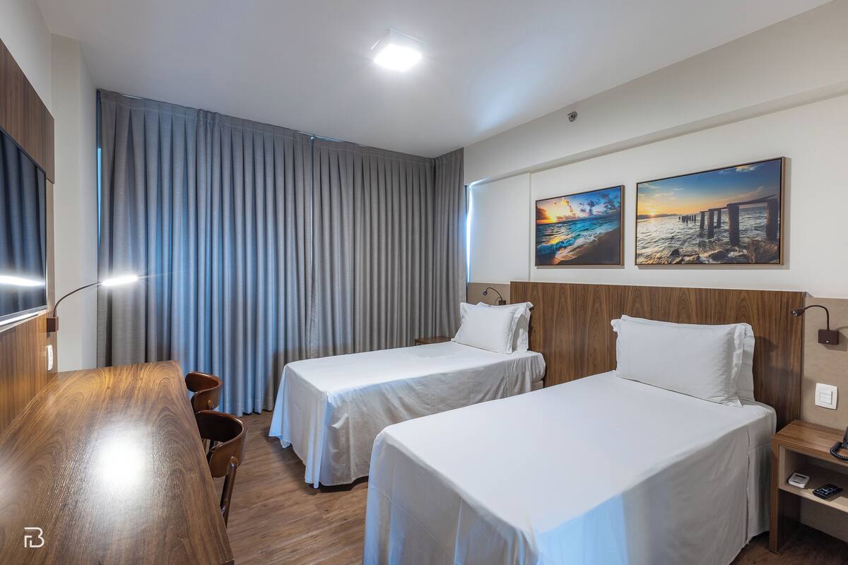 A hotel room with two single beds, a wooden desk, a chair, and a nightstand with a lamp.