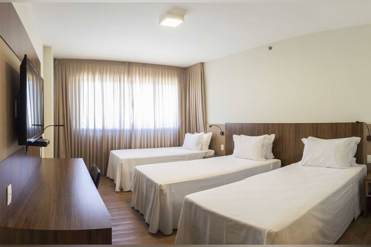A hotel room with two single beds, a TV mounted on the wall, a desk, and a large window with curtains.