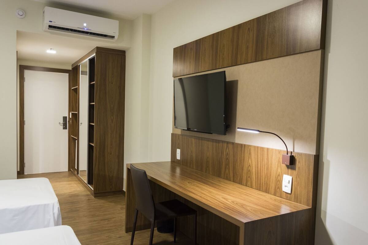 A hotel room with a wooden desk, a television mounted on the wall, a wardrobe, and a bed.