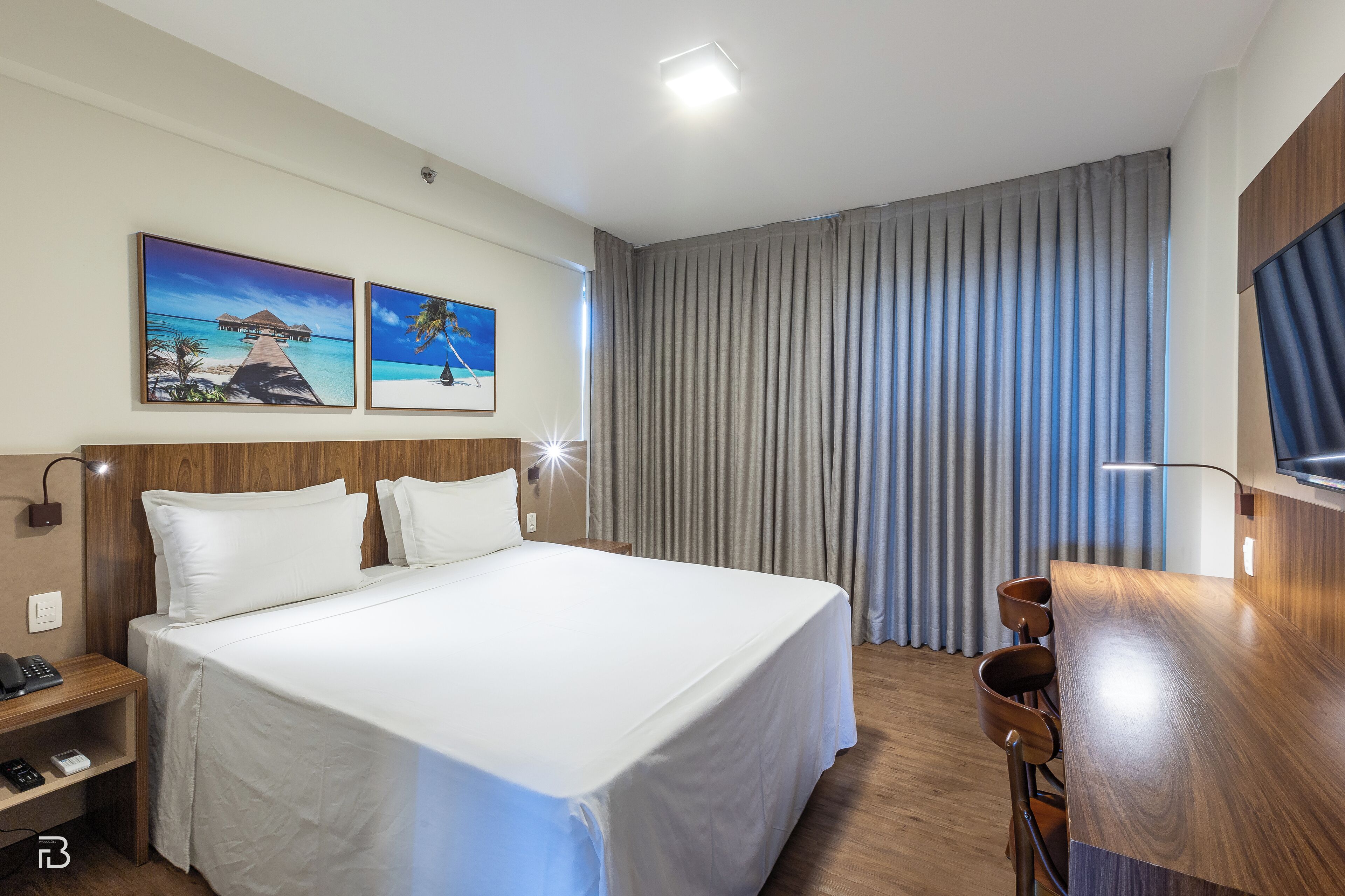 A hotel room with a large bed, a desk with a chair, a telephone, and two paintings on the wall.