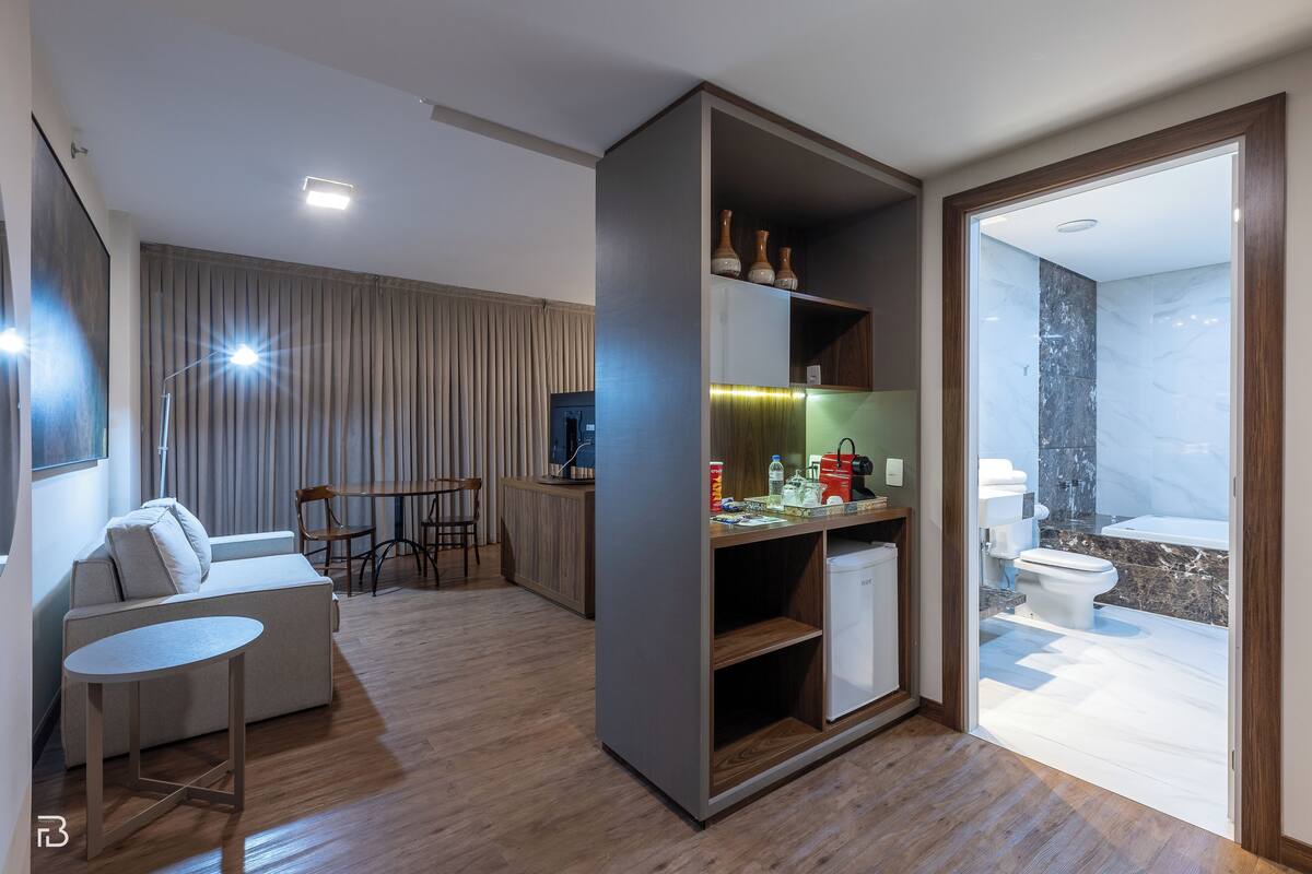 A modern living room with a sofa, dining table, and chairs. There is a kitchen area with a refrigerator, sink, and cabinets. A bathroom with a toilet and shower is visible through an open door.