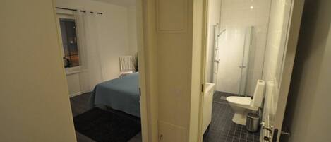 A bedroom with a bed and a small table, a bathroom with a toilet and a shower, and a door leading to another room.