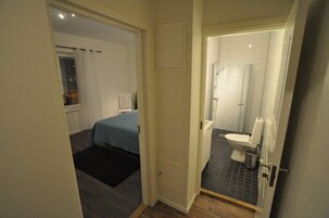 Un dormitorio con cama y una mesita, un baño con inodoro y ducha, y una puerta que da a otra habitación.