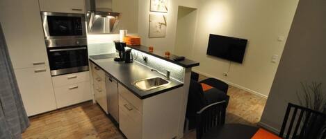 Una cocina compacta con electrodomésticos integrados, un comedor con televisor y un pequeño rincón de descanso.