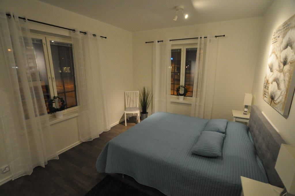 Un dormitorio con una cama grande, dos ventanas con cortinas, una silla y una mesita de noche con lámpara.