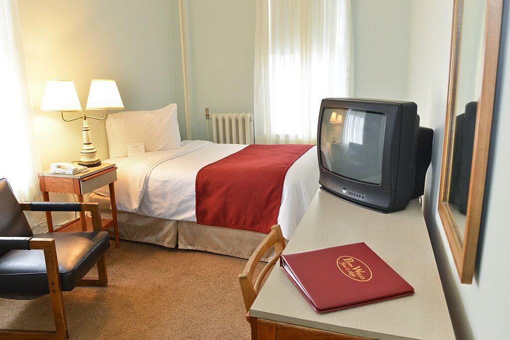 A hotel room with a bed, a desk with a television, a chair, a lamp, and a red folder on the desk.