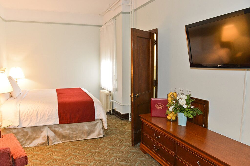 A hotel room with a bed, a TV mounted on the wall, a wooden dresser, and a desk with a lamp.