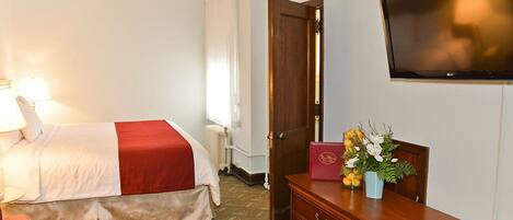 A hotel room with a bed, a TV mounted on the wall, a wooden dresser, and a desk with a lamp.