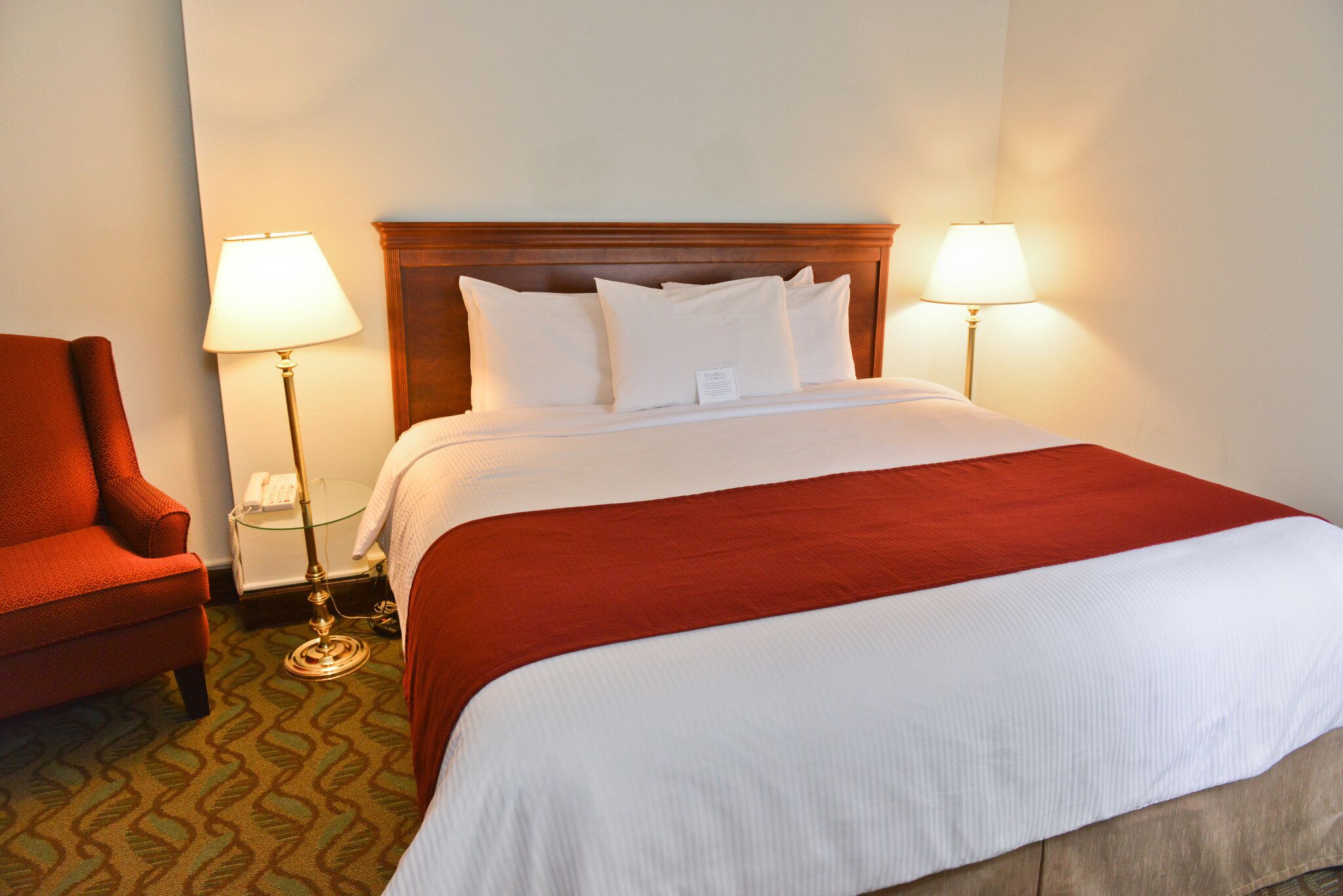 Una habitación de hotel con una cama grande, dos lámparas de noche, un sillón rojo y una alfombra estampada.