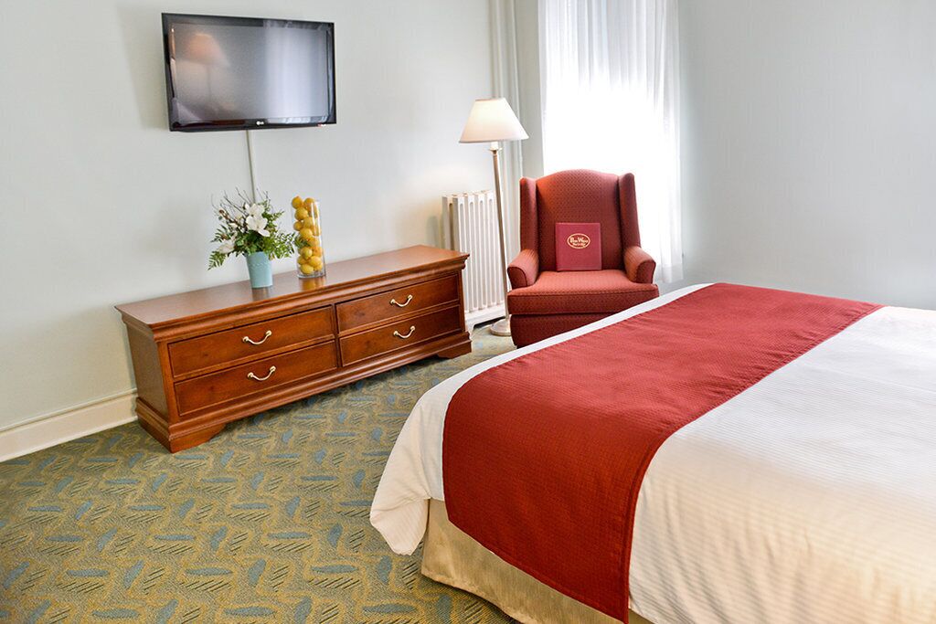 Une chambre d’hôtel avec un lit, une commode en bois, un fauteuil rouge, une télévision et une lampe.