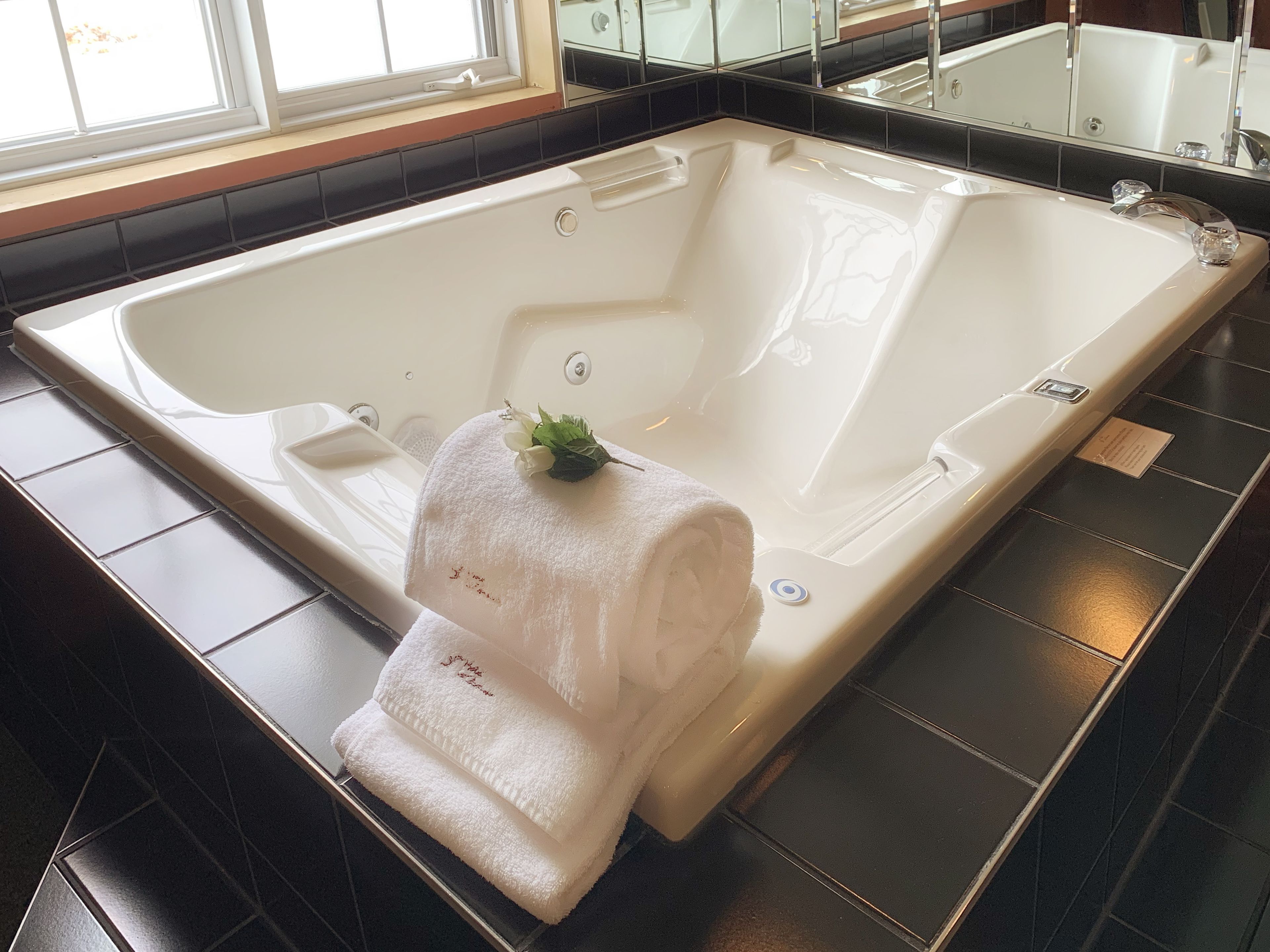 A large, white, freestanding bathtub with a single white towel and a small plant on it, set on a dark tiled floor.