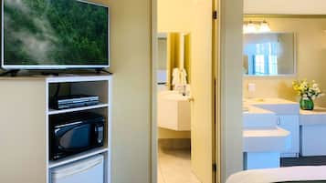 A hotel room with a television on a shelf, a microwave, a bed, a bathroom with a sink and mirror, and a door leading to another room.