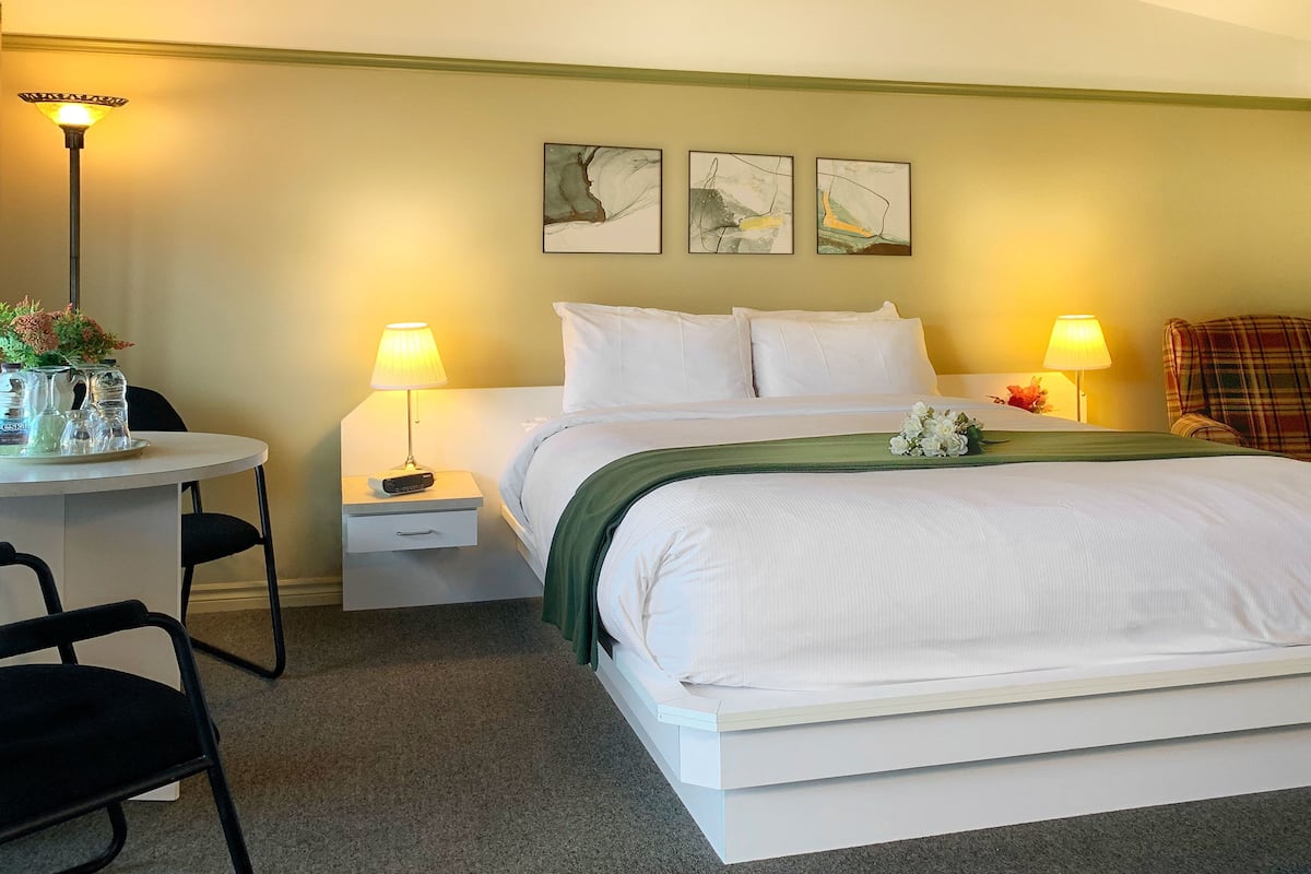 Habitación de hotel ordenada con una cama, dos lámparas de noche, una mesita con una lámpara, una silla y una pared decorada con tres cuadros.