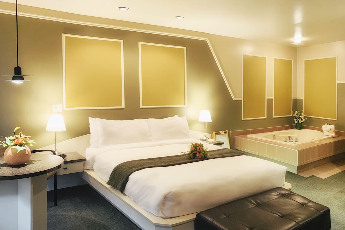 Habitación de hotel con una cama grande, lámparas de noche, una mesita con un jarrón, un reposapiés de cuero negro y un baño con bañera y espejo.