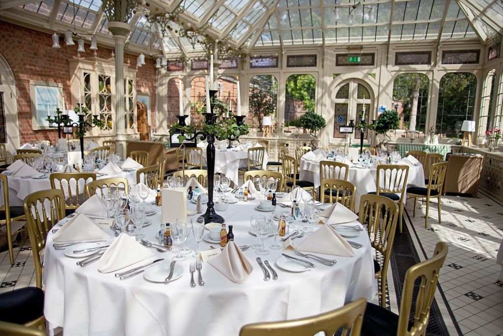A conservatory with round tables set for dining, featuring gold chairs and white tablecloths.