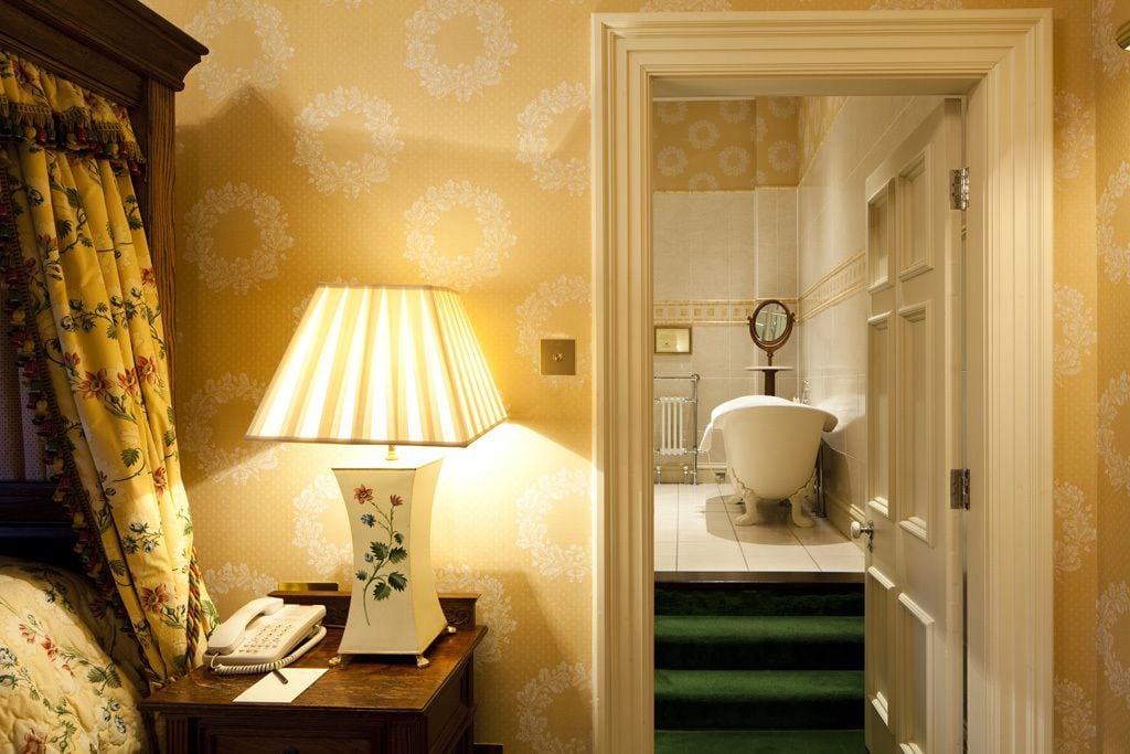 A hotel room with a bed, a bedside table with a lamp, a telephone, and a floral curtain.