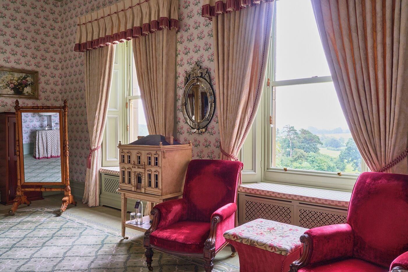 A room with floral wallpaper, a large mirror, a red velvet chair, and a view of greenery outside.