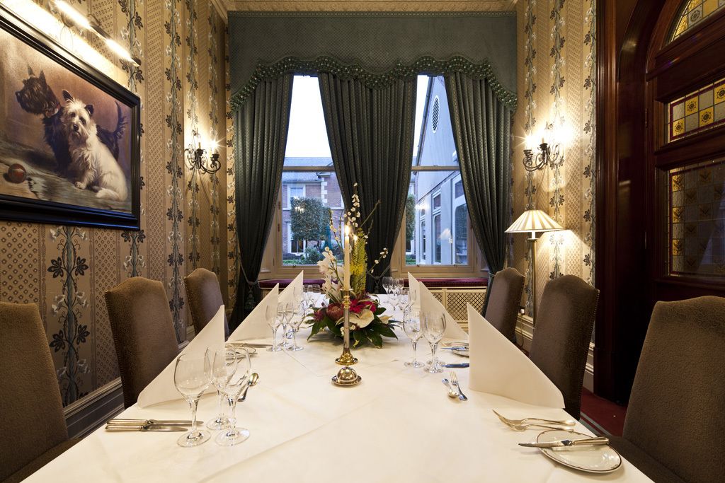 Un comedor formal con una larga mesa dispuesta para una comida, flanqueada por sillas y una ventana con cortinas.