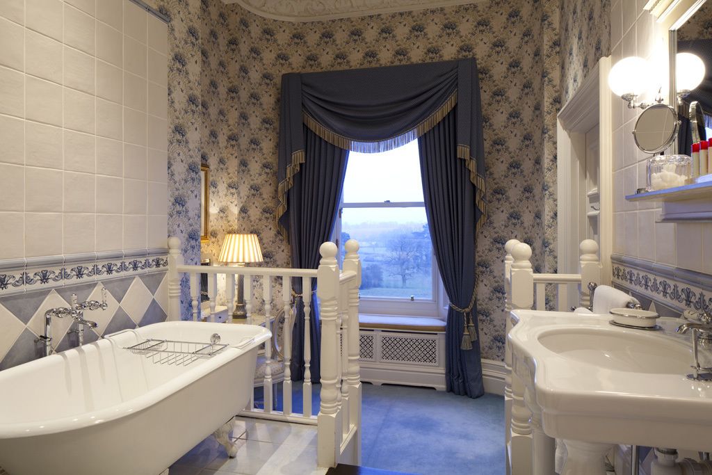 A bathroom with a bathtub, two sinks, a window with curtains, and a mirror with lights.