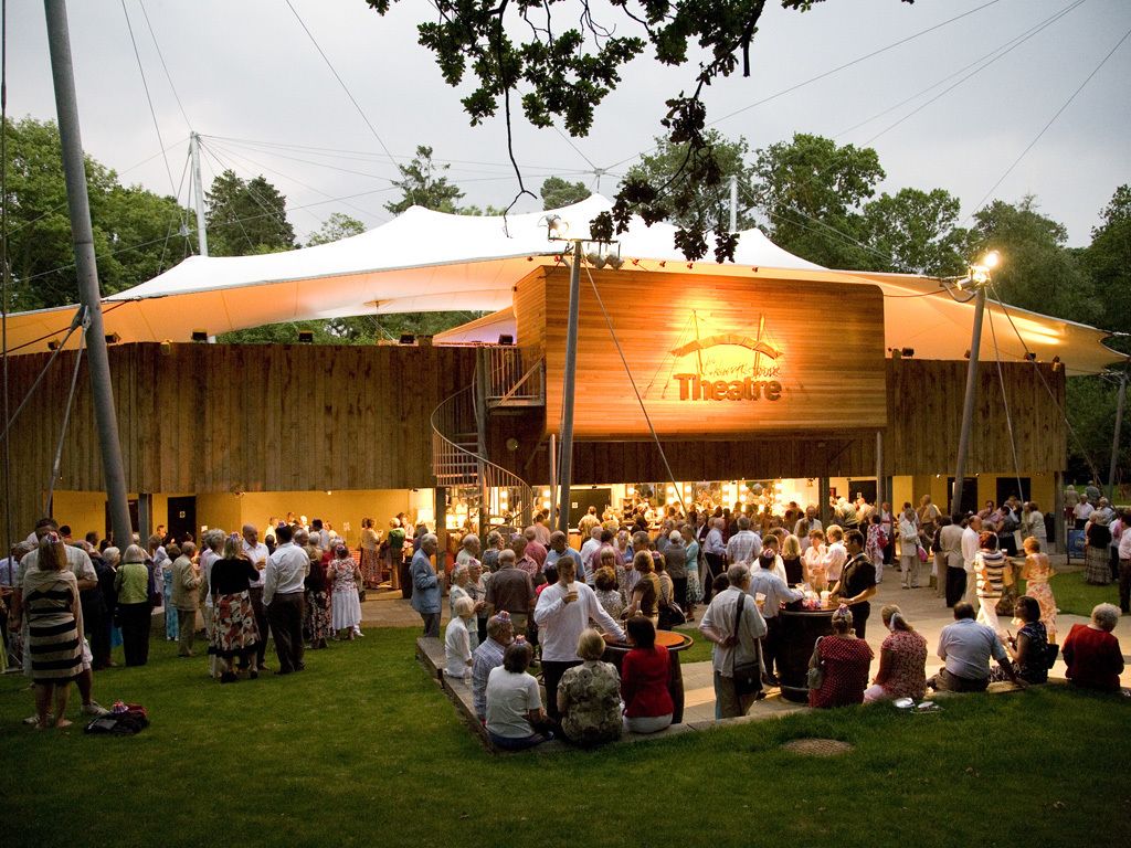 An outdoor theatre with a wooden facade and a large audience gathered under a canopy.