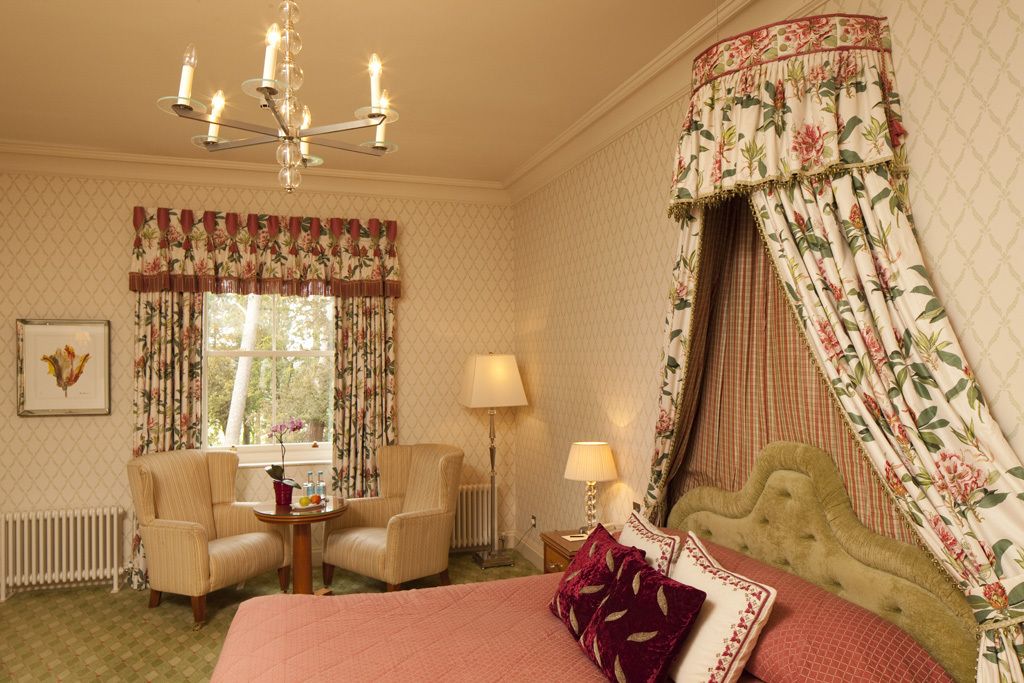 A hotel room with a bed, two armchairs, a small table, a lamp, and a chandelier.