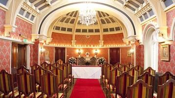 A grand hall with rows of wooden chairs, a red carpet, and ornate ceiling details.