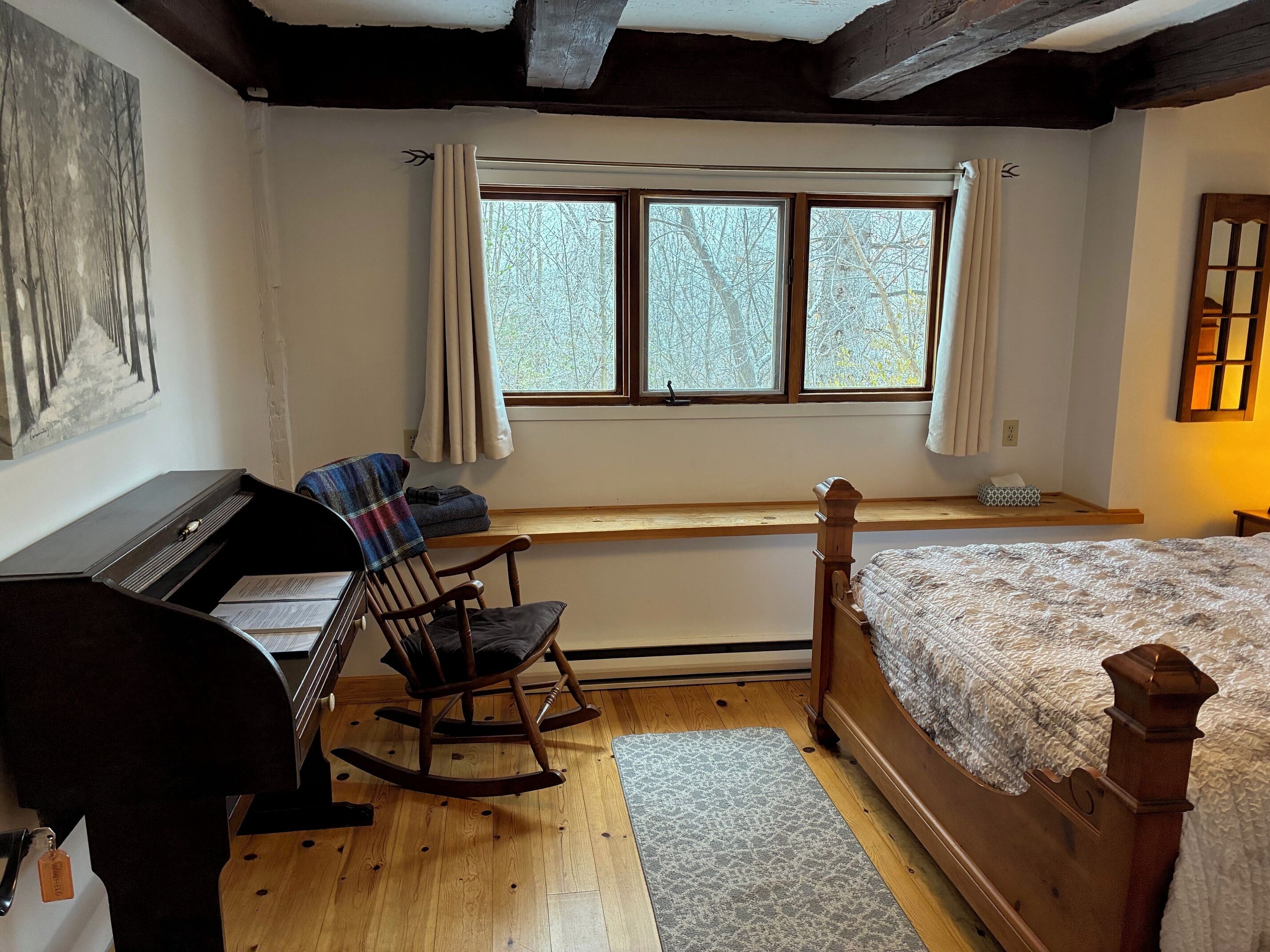 A bedroom with a wooden bed, a rocking chair, a desk with a chair, a window with curtains, and a painting on the wall.