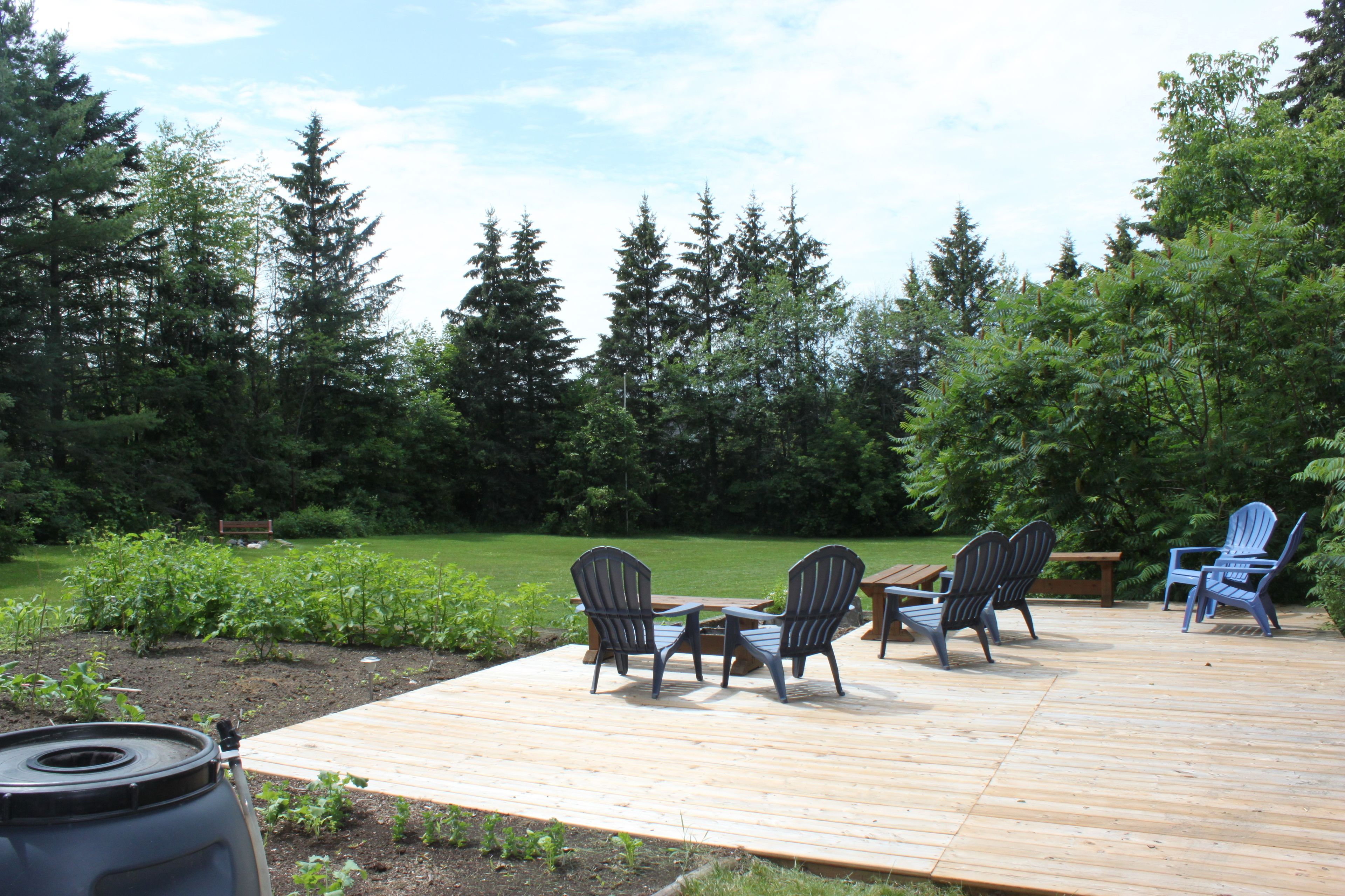 Eine Holzterrasse mit Adirondack-Stühlen und einem Tisch, umgeben von einem Garten und hohen Bäumen.