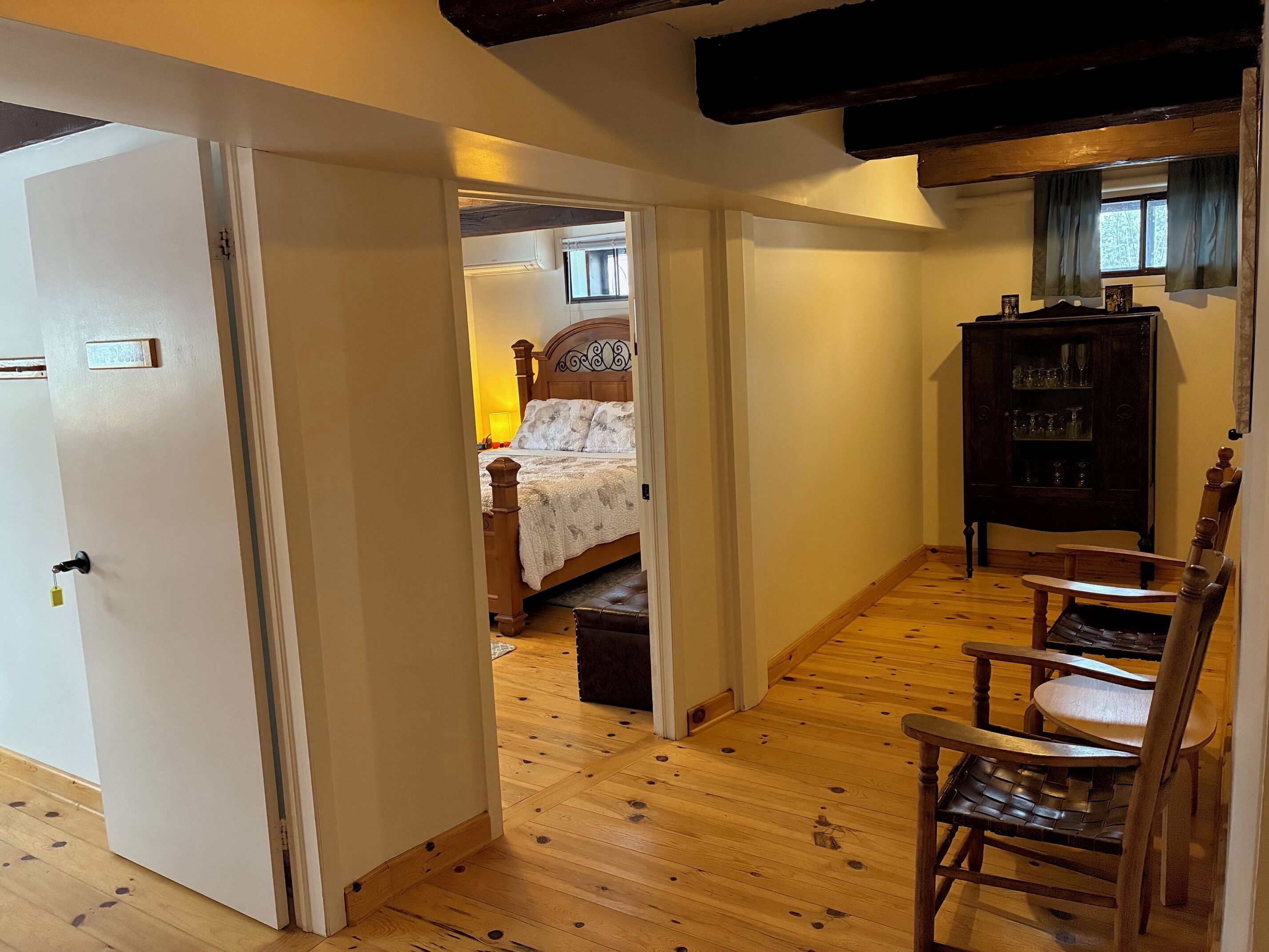 A narrow hallway with wooden flooring leading to a bedroom with a bed and a nightstand. There is a wooden chair and a cabinet with glassware in the room.