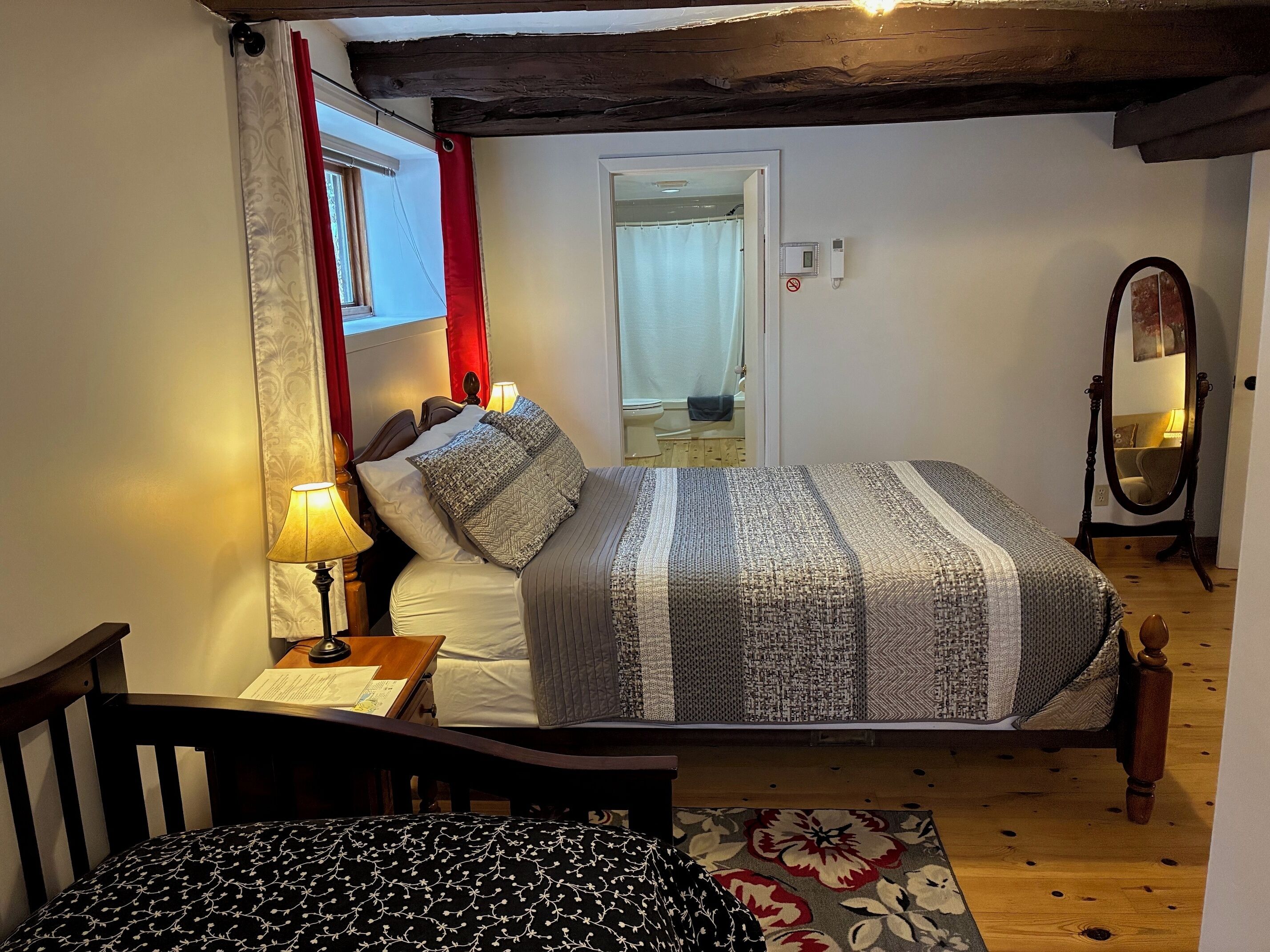 Un dormitorio con una cama, una silla de madera, un escritorio con una lámpara, un espejo y una puerta que da a un baño.
