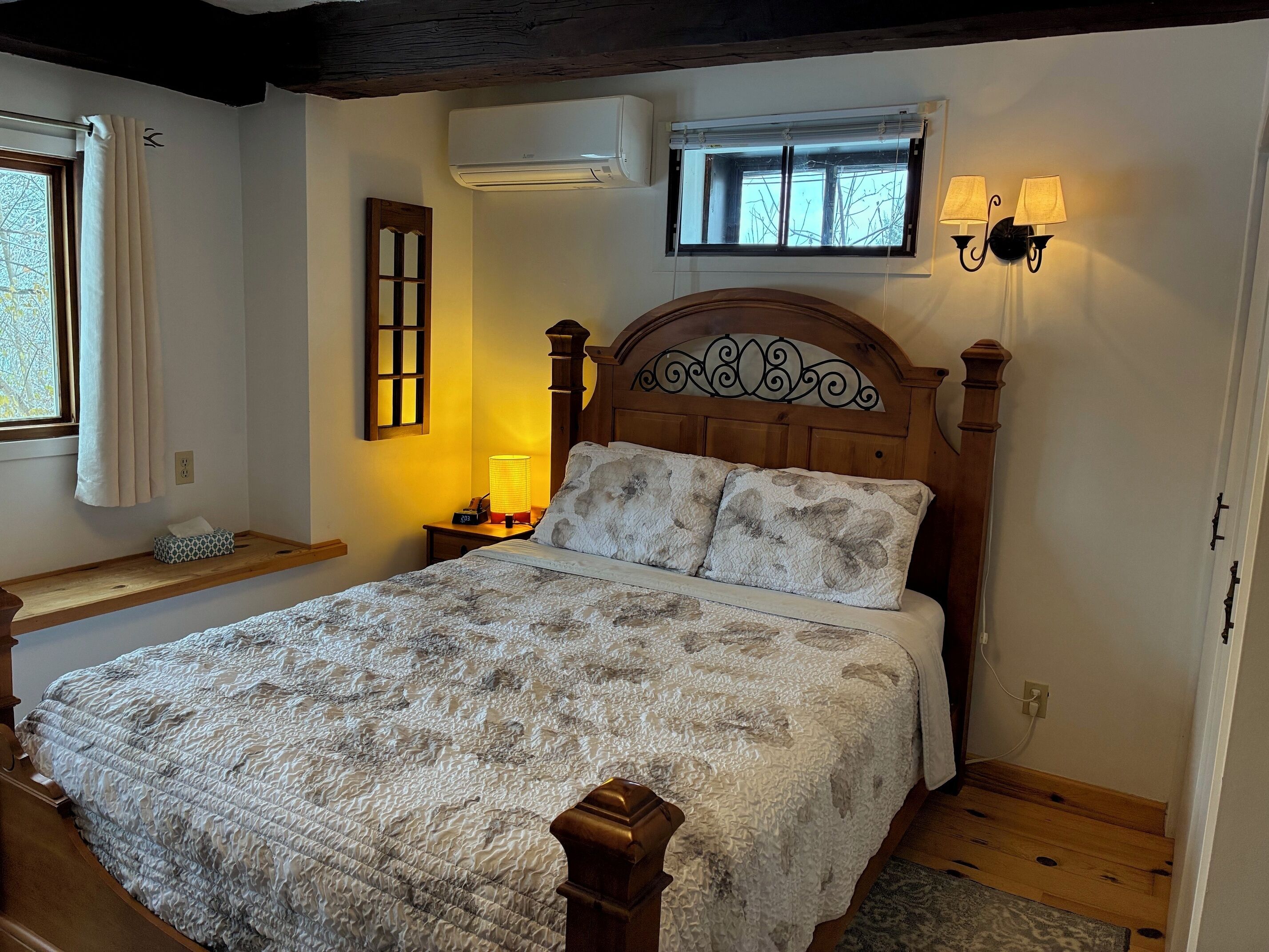 A bedroom with a wooden bed, a nightstand with a lamp, an air conditioner, a window with curtains, and a wall-mounted light fixture.