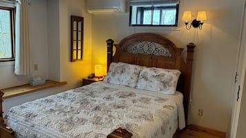 A bedroom with a wooden bed, a nightstand with a lamp, an air conditioner, a window with curtains, and a wall-mounted light fixture.