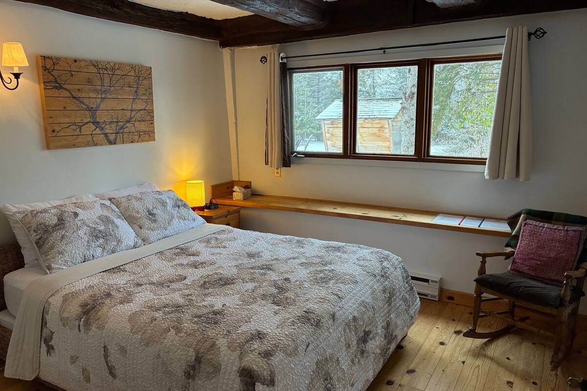A bedroom with a large bed, a bedside table with a lamp, a window with curtains, a wooden bench, and a chair.
