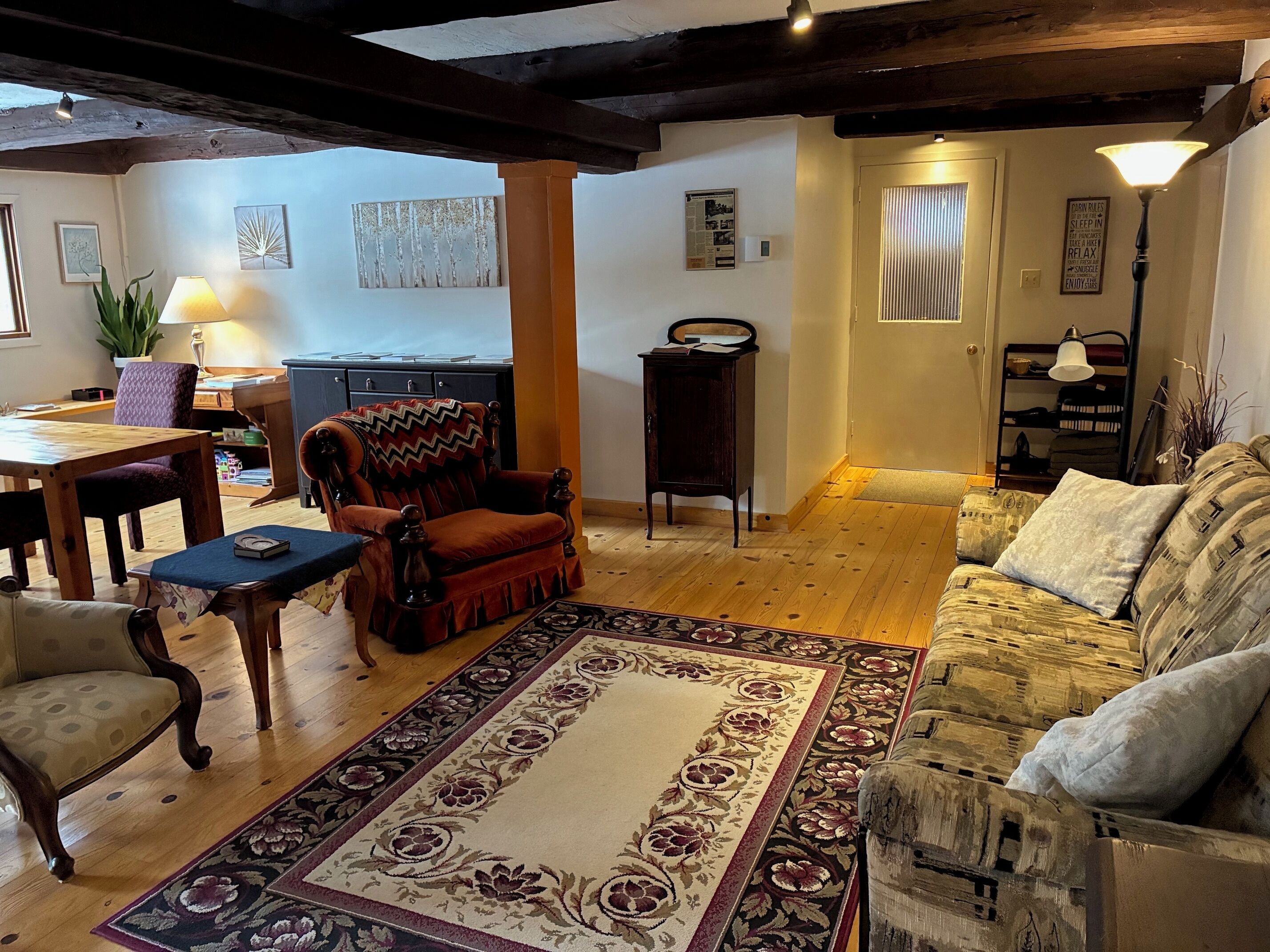 A cozy living room with a wooden floor, a patterned rug, a red armchair, a sofa, and a dining area with a table and chairs.