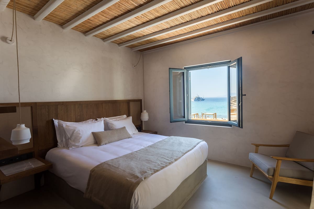 A bedroom with a large bed, wooden headboard, a nightstand, a chair, and a window with a view of the sea.