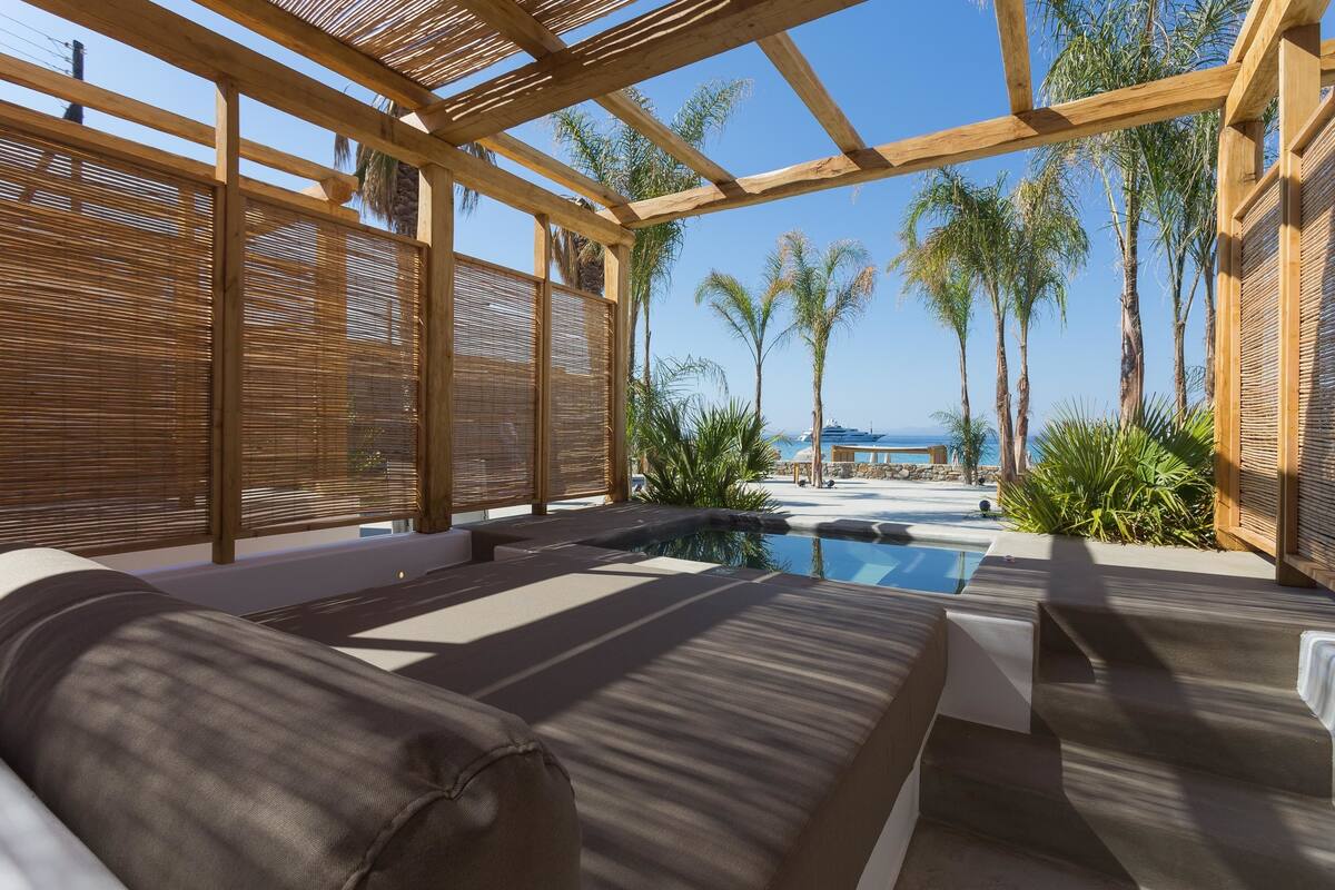 A sunlit outdoor lounge area with a pool, wooden pergola, and palm trees.